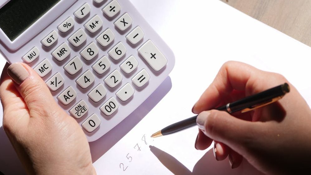 Hands using a calculator and writing numbers on paper under sunlight. Ideal for finance and education themes.